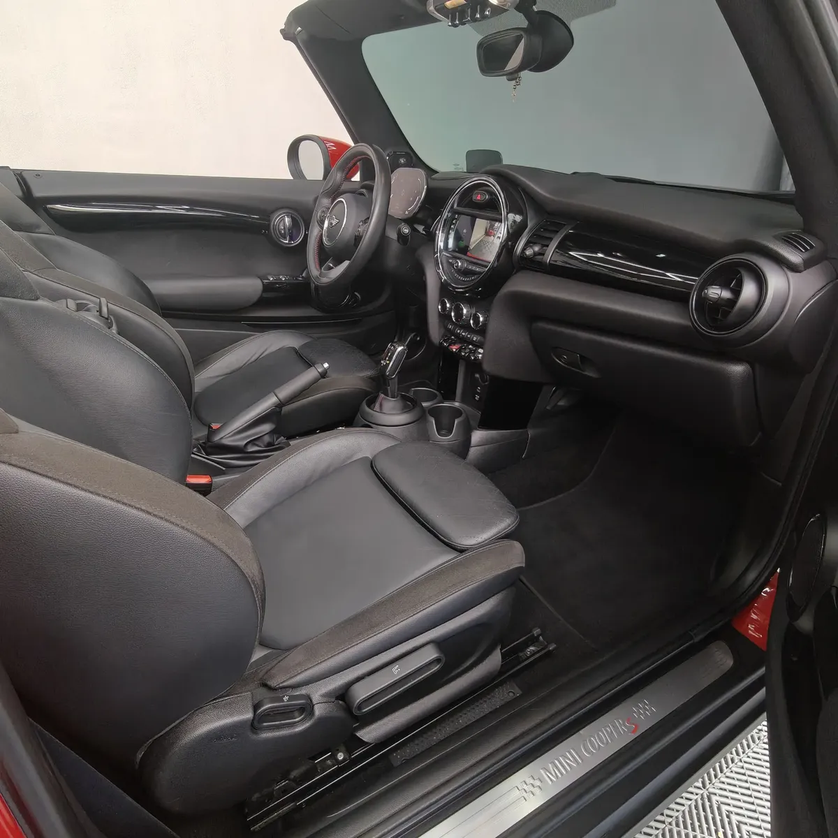 Vista lateral izquierda del habitáculo interior de un Mini Cooper S Convertible Hot Chili 2021 seminuevo, mostrando los asientos deportivos de piel negros y el tablero de instrumentos con la capota abierta, en Namacar Automotriz.