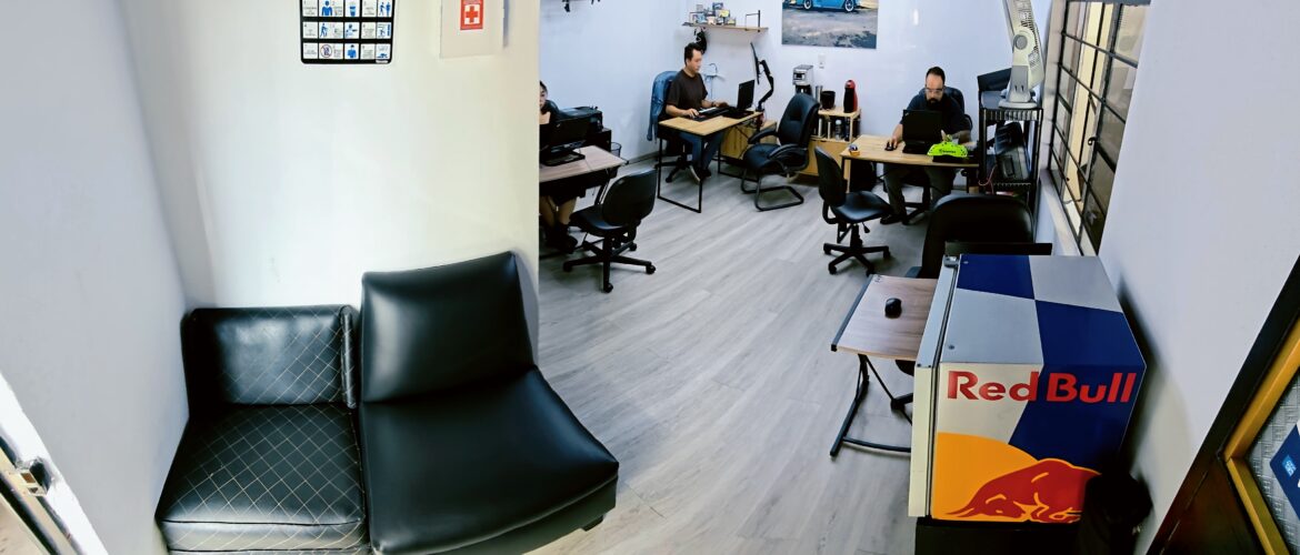 Oficina moderna con escritorios, personal trabajando en computadoras, señalización de seguridad y decoración con una camioneta clásica azul.