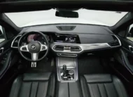 Vista interior cenital desde el techo abierto que muestra la cabina delantera con tablero digital, volante multifunción y consola central de una SUV de lujo BMW X5 2021 seminueva en color blanco.