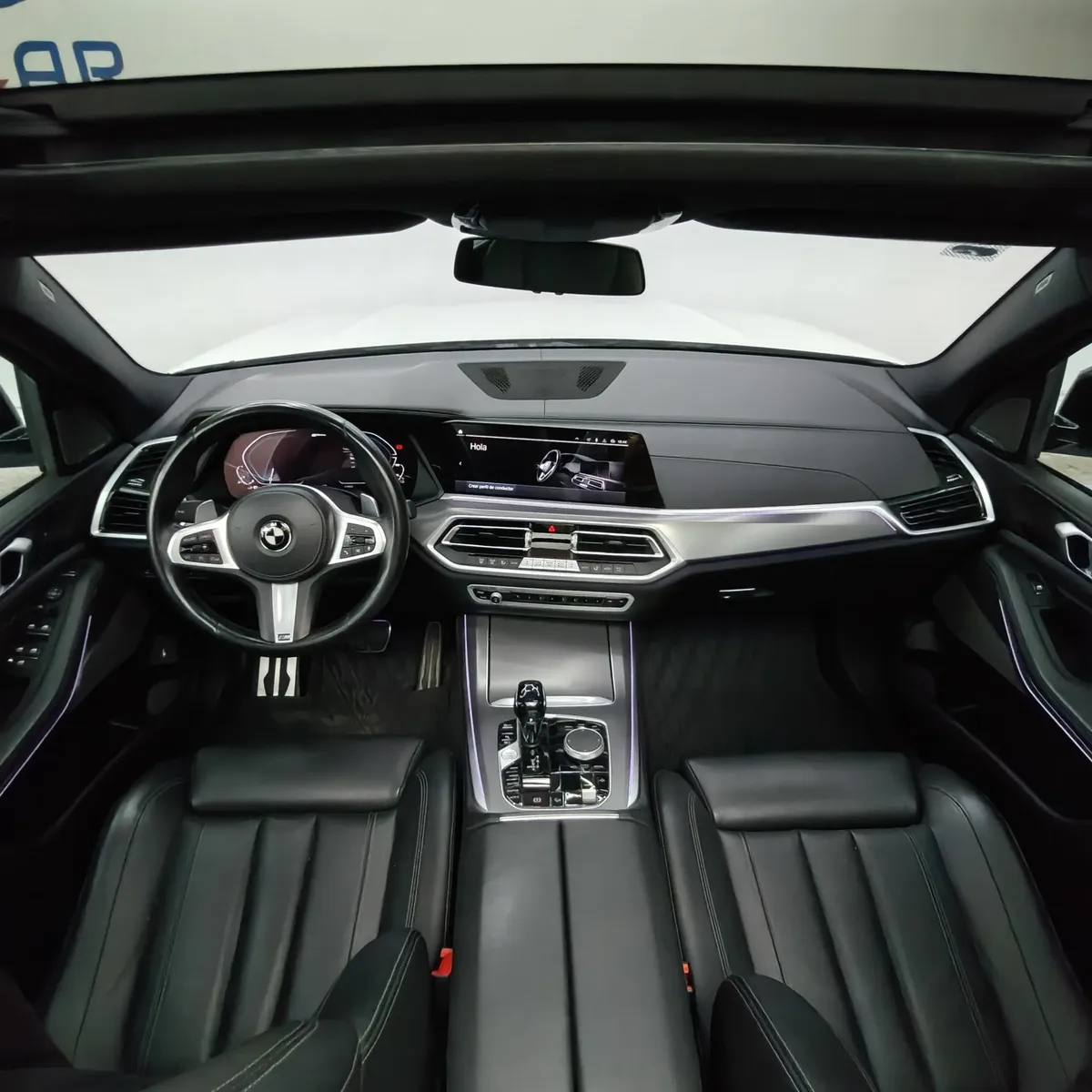 Vista interior cenital desde el techo abierto que muestra la cabina delantera con tablero digital, volante multifunción y consola central de una SUV de lujo BMW X5 2021 seminueva en color blanco.