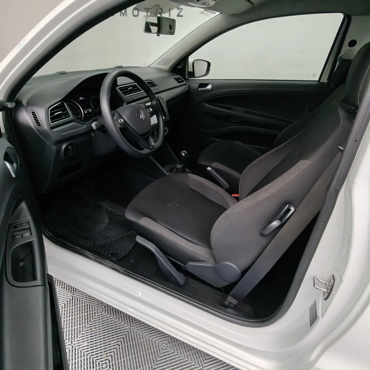 Vista interior desde la puerta del conductor abierta que muestra el asiento de tela del piloto, el volante y el tablero de una camioneta pick-up Volkswagen Saveiro 1.6 Robust MT año 2025 seminueva en ángulo oblicuo izquierdo.