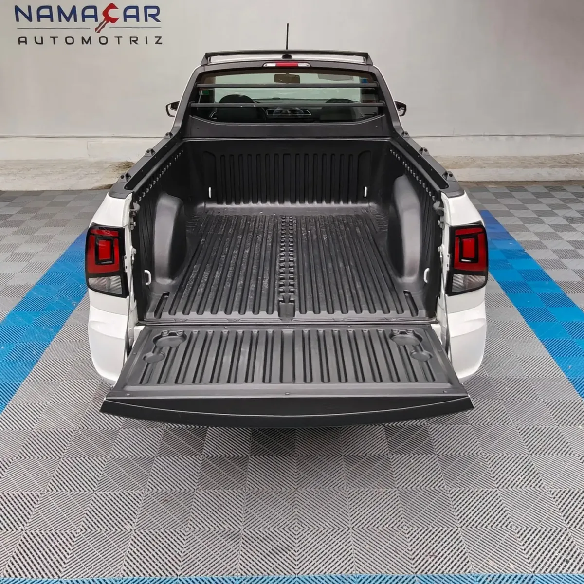 Vista exterior trasera de la batea de carga abierta con recubrimiento plástico protector (bedliner) de una camioneta pick-up Volkswagen Saveiro 1.6 Robust MT año 2025 seminueva en color blanco, en Namacar Automotriz.