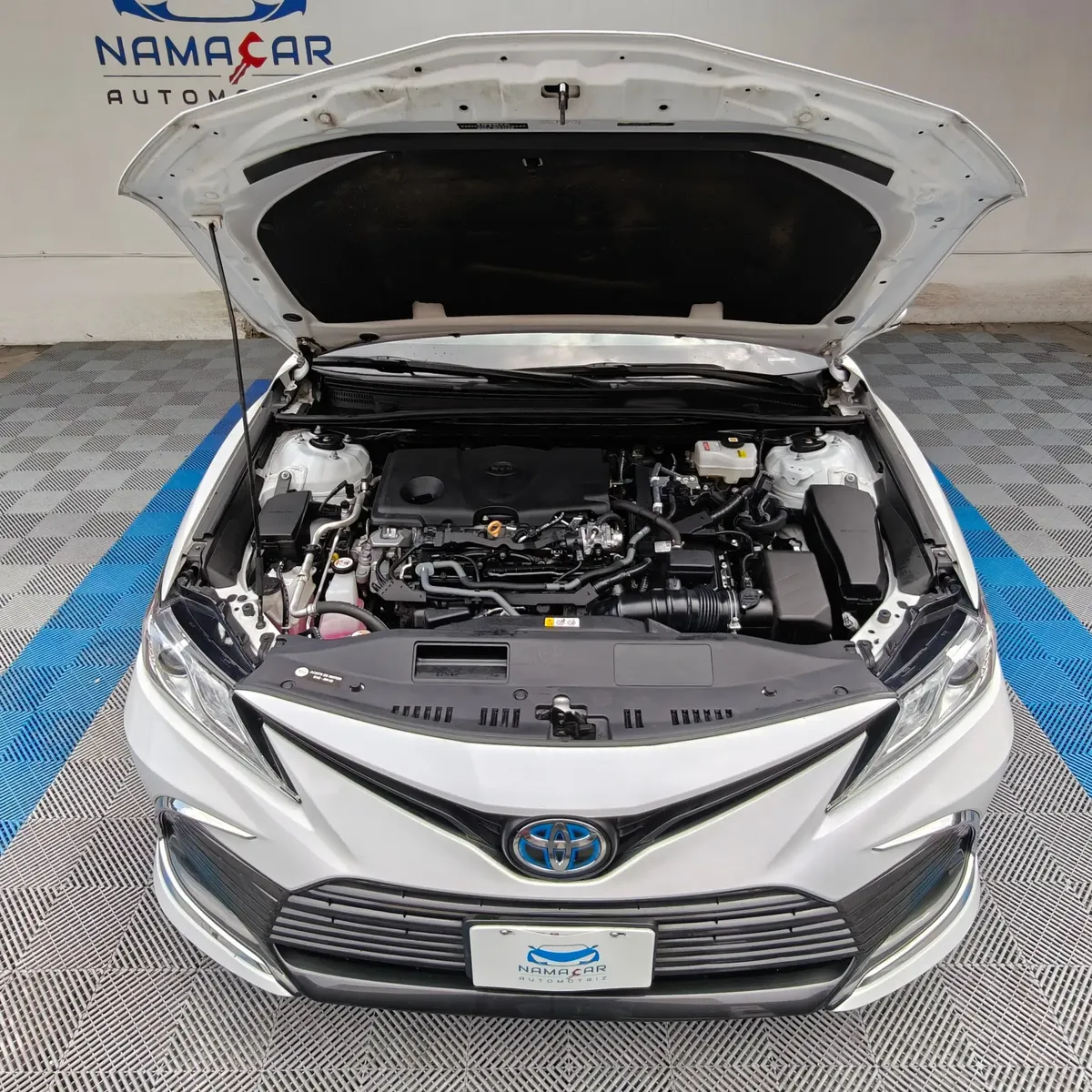 Vista exterior frontal en picada superior con el cofre abierto que muestra el motor 2.5 L híbrido de un auto sedán Toyota Camry 2.5 HEV XLE año 2024 seminuevo en color blanco, en Namacar Automotriz.