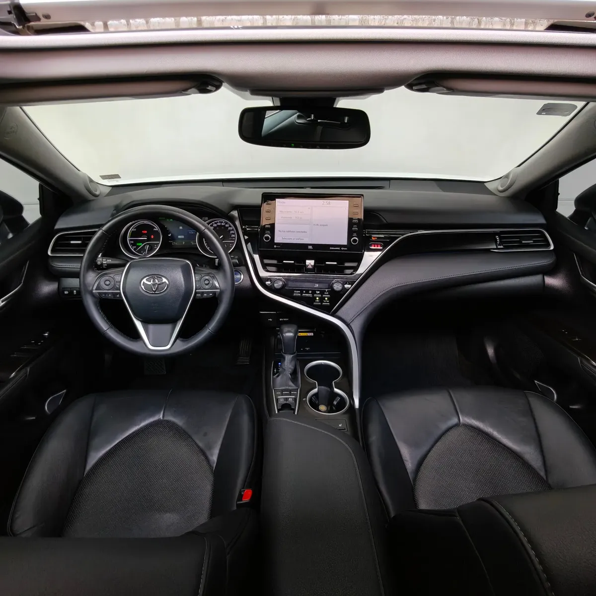 Vista interior cenital desde arriba que muestra la cabina, el tablero, la pantalla multimedia flotante, el volante multifunción y los asientos delanteros en piel negra de un auto sedán híbrido Toyota Camry 2.5 HEV XLE año 2024 seminuevo.