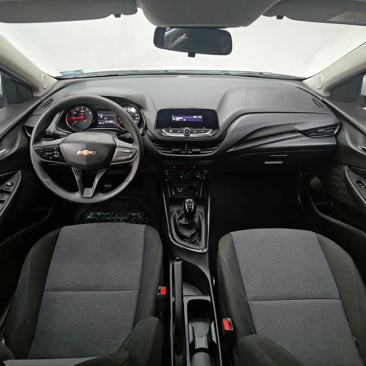 Vista interior cenital desde arriba que muestra la cabina, el tablero, el sistema de audio, el volante multifunción y los asientos delanteros en tela de un auto sedán Chevrolet Onix 1.3 LS manual año 2024 seminuevo.