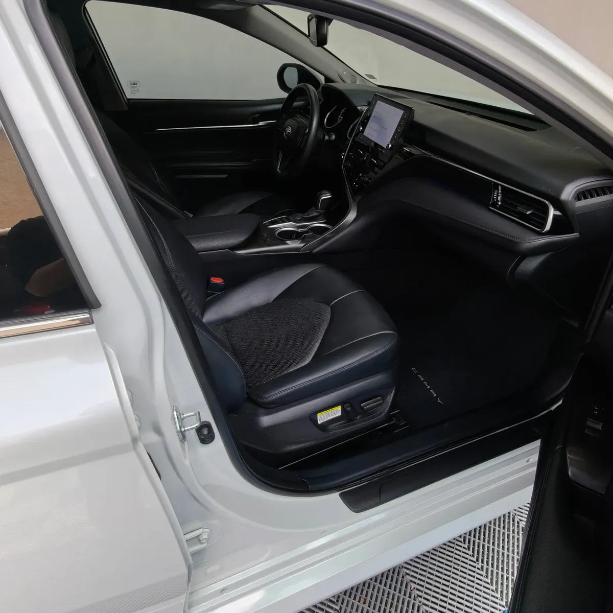 Vista interior desde la puerta del copiloto abierta que muestra el asiento de piel, la guantera, el tablero y la consola central de un auto sedán híbrido Toyota Camry 2.5 HEV XLE año 2024 seminuevo en ángulo oblicuo derecho.