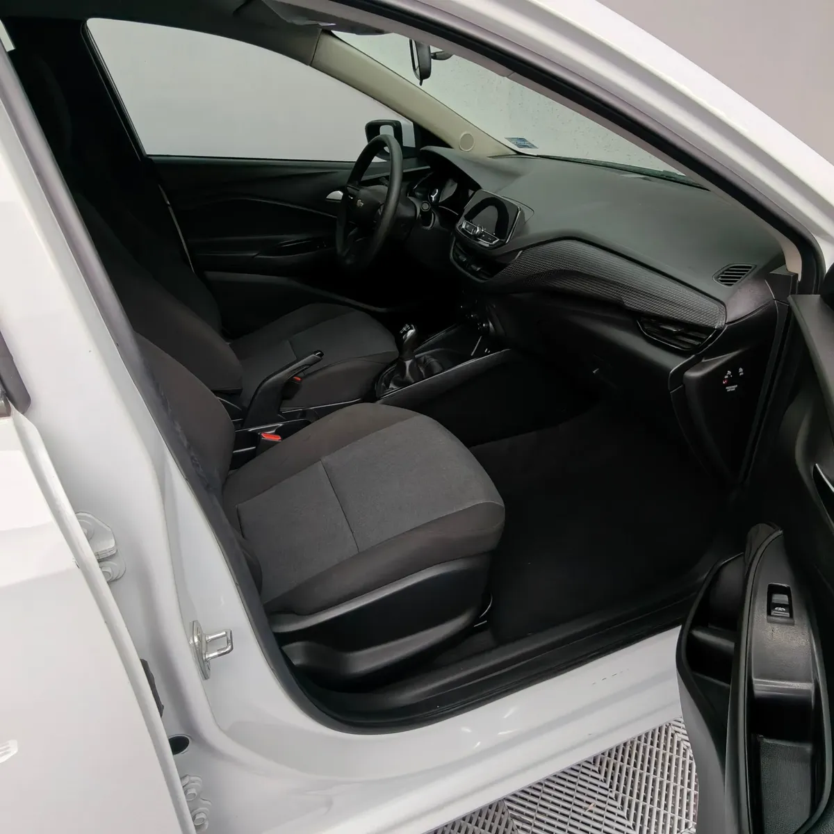Vista interior desde la puerta del copiloto abierta que muestra el asiento en tela del acompañante, la guantera, el tablero y la consola central de un auto sedán Chevrolet Onix 1.3 LS manual año 2024 seminuevo en ángulo oblicuo derecho.