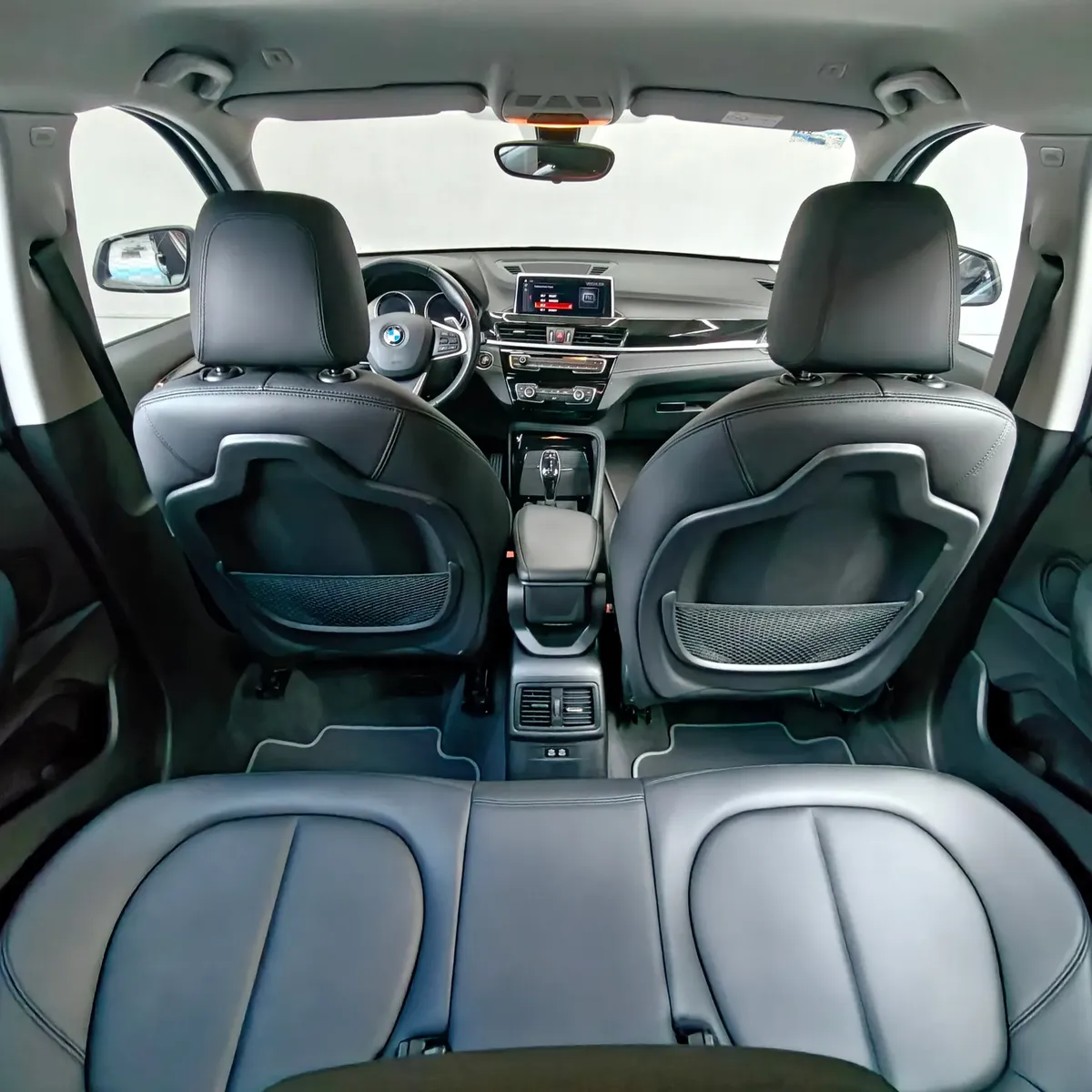 Vista interior amplia desde la cajuela y zona posterior enfocando la banca de asientos traseros en piel negra, respaldos delanteros con redes portaobjetos, consola central trasera y vista hacia la cabina frontal de una camioneta SUV de lujo BMW X1 2.0 sDrive20i sDrive 20iA X Line transmisión automática año 2022 seminueva.