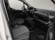 Vista interior desde la puerta del copiloto abierta que muestra el asiento delantero derecho en tela oscura, la consola central con palanca de velocidades integrada, el tablero con compartimentos portaobjetos y la cabina de una camioneta comercial Peugeot Partner 1.6 HDi Maxi transmisión manual año 2021 seminueva en ángulo oblicuo derecho.