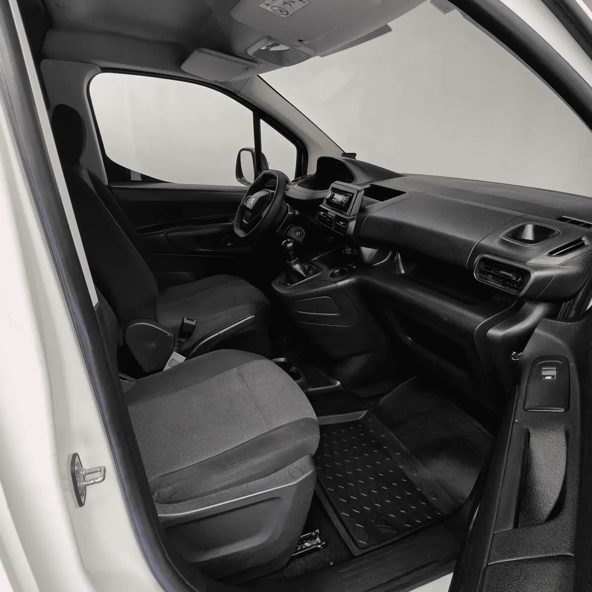 Vista interior desde la puerta del copiloto abierta que muestra el asiento delantero derecho en tela oscura, la consola central con palanca de velocidades integrada, el tablero con compartimentos portaobjetos y la cabina de una camioneta comercial Peugeot Partner 1.6 HDi Maxi transmisión manual año 2021 seminueva en ángulo oblicuo derecho.