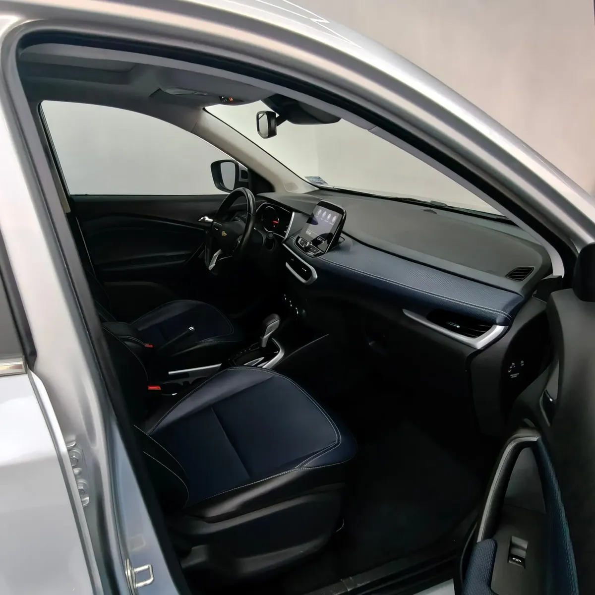 Vista interior desde la puerta del copiloto abierta que muestra el asiento delantero derecho forrado en piel bitono azul y negro, el tablero con acabados en piel, la pantalla de infoentretenimiento y la consola central de una camioneta SUV Chevrolet Tracker 1.2 Premier Turbo transmisión automática año 2023 seminueva en ángulo oblicuo derecho.
