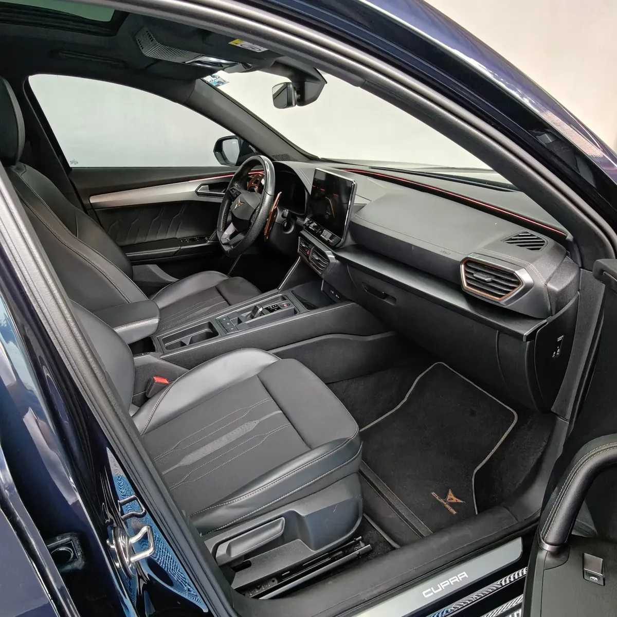 Vista interior desde la puerta del copiloto abierta que muestra el asiento delantero derecho, el tapete con logotipo Cupra, la consola central, la pantalla táctil y el tablero de una SUV Crossover deportiva Cupra Formentor 2.0 Mid 190 HP 2WD DSG7 año 2022 seminueva en ángulo oblicuo derecho.
