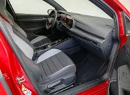 Vista interior desde la puerta del copiloto abierta que muestra el asiento delantero derecho con tapicería deportiva bitono y costuras rojas, la consola central, la pantalla táctil y el tablero de un hatchback deportivo Volkswagen Golf GTI 2.0 241 HP DSG7 año 2024 seminuevo en ángulo oblicuo derecho.