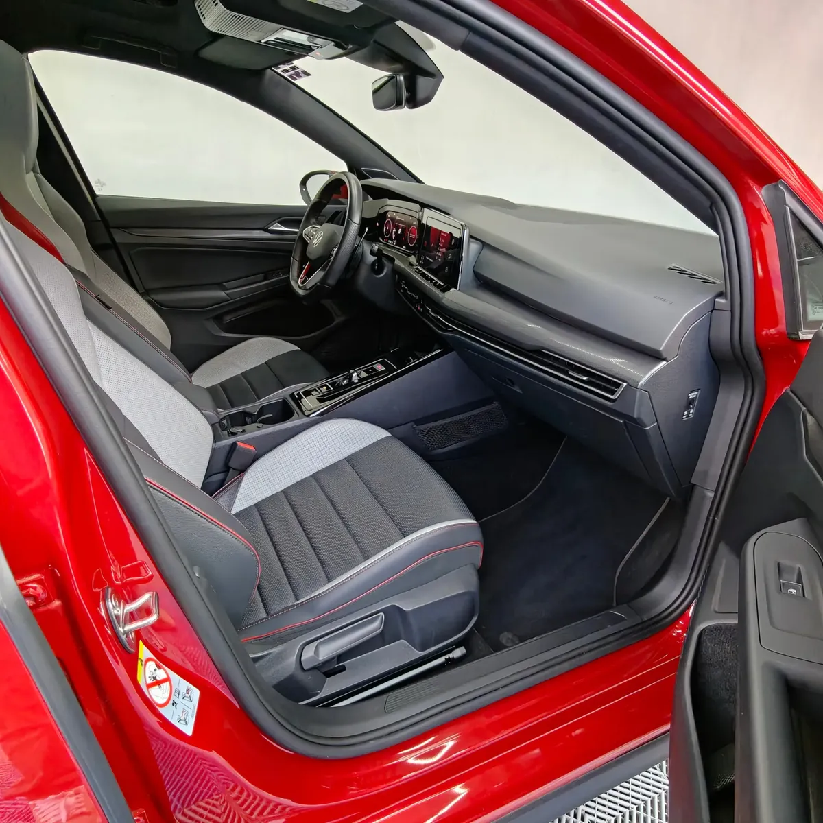 Vista interior desde la puerta del copiloto abierta que muestra el asiento delantero derecho con tapicería deportiva bitono y costuras rojas, la consola central, la pantalla táctil y el tablero de un hatchback deportivo Volkswagen Golf GTI 2.0 241 HP DSG7 año 2024 seminuevo en ángulo oblicuo derecho.