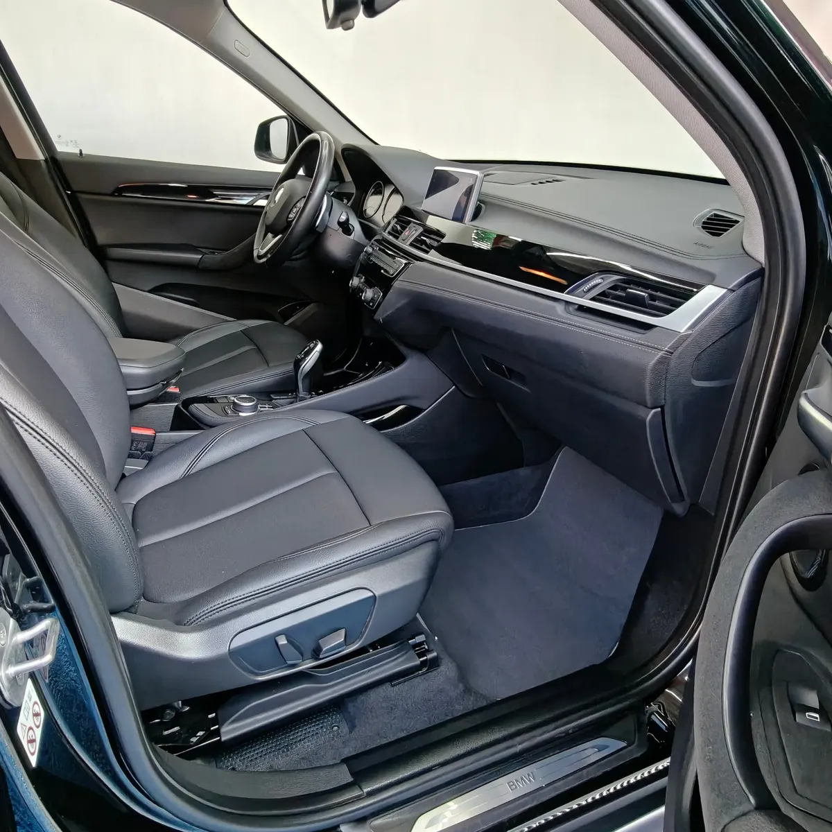 Vista interior desde la puerta del copiloto abierta que muestra el asiento delantero derecho en piel negra con ajustes eléctricos, la consola central, la pantalla táctil y el tablero de una camioneta SUV de lujo BMW X1 2.0 sDrive20i sDrive 20iA X Line transmisión automática año 2022 seminueva en ángulo oblicuo derecho.