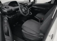 Vista interior desde la puerta del conductor abierta que muestra el puesto de mando ergonómico i-Cockpit, el asiento del piloto en tela oscura con ajustes manuales, el volante multifunción compacto, los pedales de control y el panel de la puerta izquierda de una camioneta comercial Peugeot Partner 1.6 HDi Maxi transmisión manual año 2021 seminueva en ángulo oblicuo izquierdo.