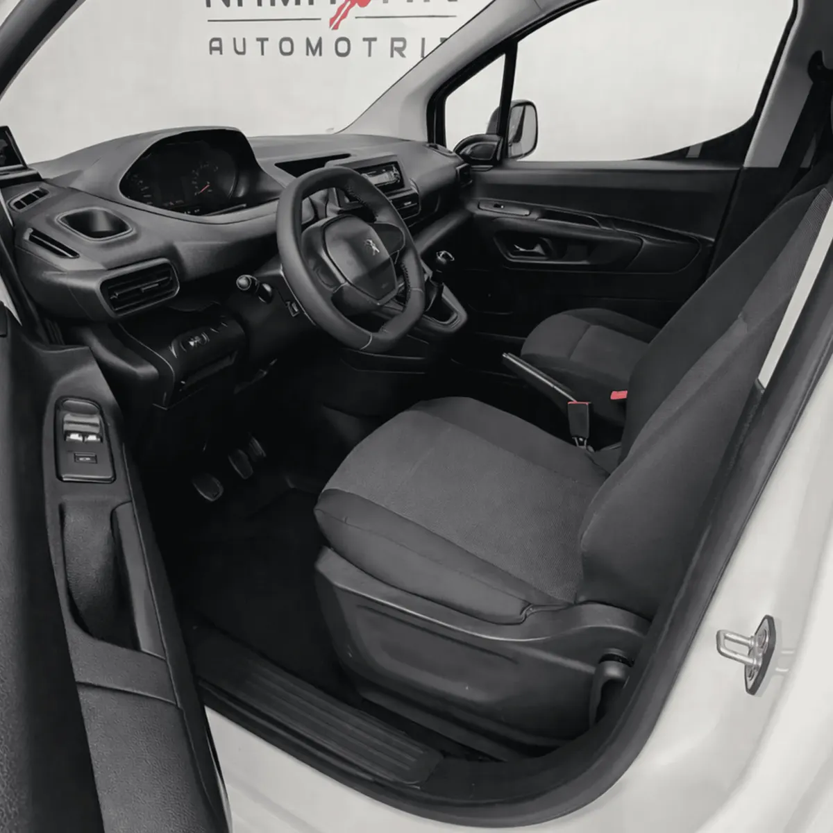 Vista interior desde la puerta del conductor abierta que muestra el puesto de mando ergonómico i-Cockpit, el asiento del piloto en tela oscura con ajustes manuales, el volante multifunción compacto, los pedales de control y el panel de la puerta izquierda de una camioneta comercial Peugeot Partner 1.6 HDi Maxi transmisión manual año 2021 seminueva en ángulo oblicuo izquierdo.