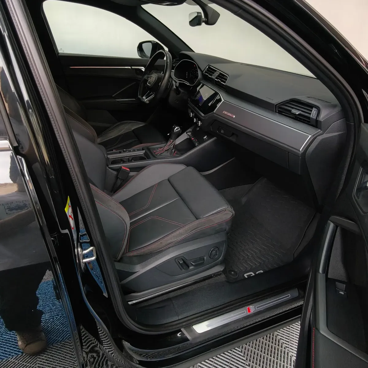 Vista interior desde la puerta del copiloto abierta que muestra el asiento deportivo delantero derecho con ajustes eléctricos forrado en piel negra con costuras rojas, el estribo de aluminio iluminado S Line, el tablero con emblema quattro y la consola central de una camioneta SUV Audi Q3 2.0 S Line 180 Hp transmisión DSG automática año 2022 seminueva en ángulo oblicuo derecho.
