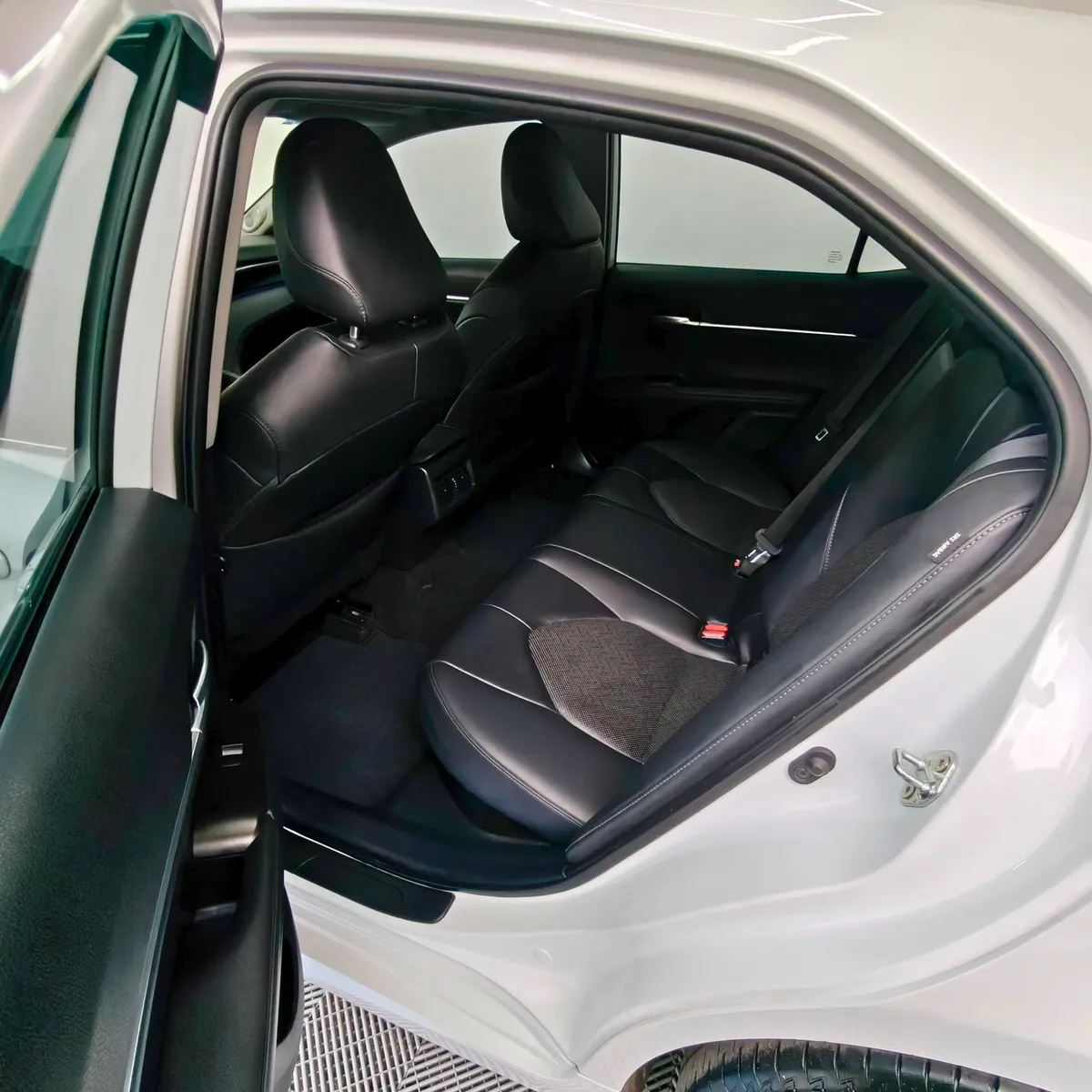 Vista interior desde la puerta trasera izquierda abierta que muestra la banca de asientos posteriores en piel negra, los respaldos delanteros y el área de pasajeros de un auto sedán híbrido Toyota Camry 2.5 HEV XLE año 2024 seminuevo en ángulo oblicuo izquierdo.