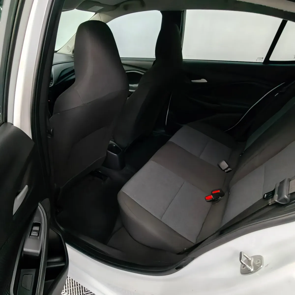 Vista interior desde la puerta trasera izquierda abierta que muestra la banca de asientos posteriores en tela gris, el espacio para piernas y los respaldos delanteros de un auto sedán Chevrolet Onix 1.3 LS manual año 2024 seminuevo en ángulo oblicuo izquierdo.