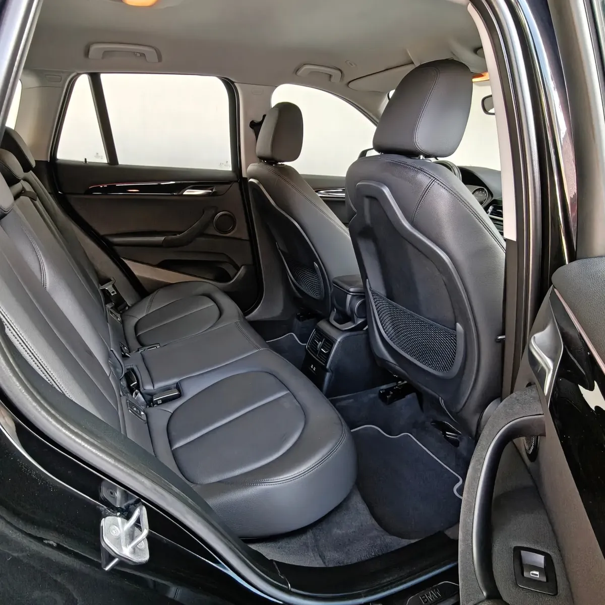Vista interior desde la puerta trasera derecha abierta que muestra la banca de asientos posteriores tapizados en piel negra, el espacio para piernas, las salidas de aire acondicionado traseras y el respaldo del asiento del copiloto de una camioneta SUV de lujo BMW X1 2.0 sDrive20i sDrive 20iA X Line transmisión automática año 2022 seminueva en ángulo oblicuo derecho.
