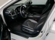 Vista interior desde la puerta del conductor abierta que muestra el asiento del piloto, el volante multifunción, el cuadro de instrumentos y el tablero de un auto sedán híbrido Toyota Camry 2.5 HEV XLE año 2024 seminuevo en ángulo oblicuo izquierdo.