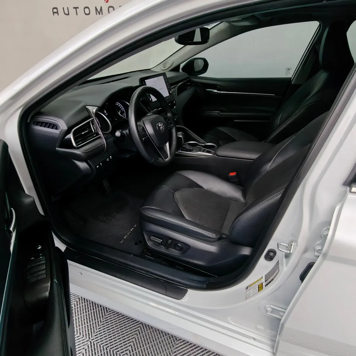 Vista interior desde la puerta del conductor abierta que muestra el asiento del piloto, el volante multifunción, el cuadro de instrumentos y el tablero de un auto sedán híbrido Toyota Camry 2.5 HEV XLE año 2024 seminuevo en ángulo oblicuo izquierdo.
