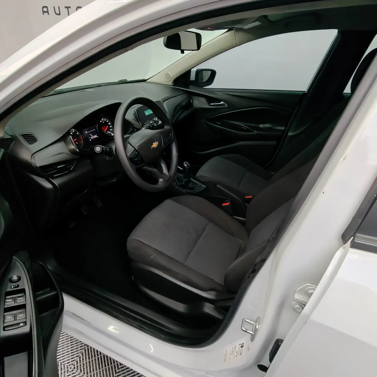 Vista interior desde la puerta del conductor abierta que muestra el asiento del piloto en tela, el volante multifunción, el cuadro de instrumentos y el tablero de un auto sedán Chevrolet Onix 1.3 LS manual año 2024 seminuevo en ángulo oblicuo izquierdo.