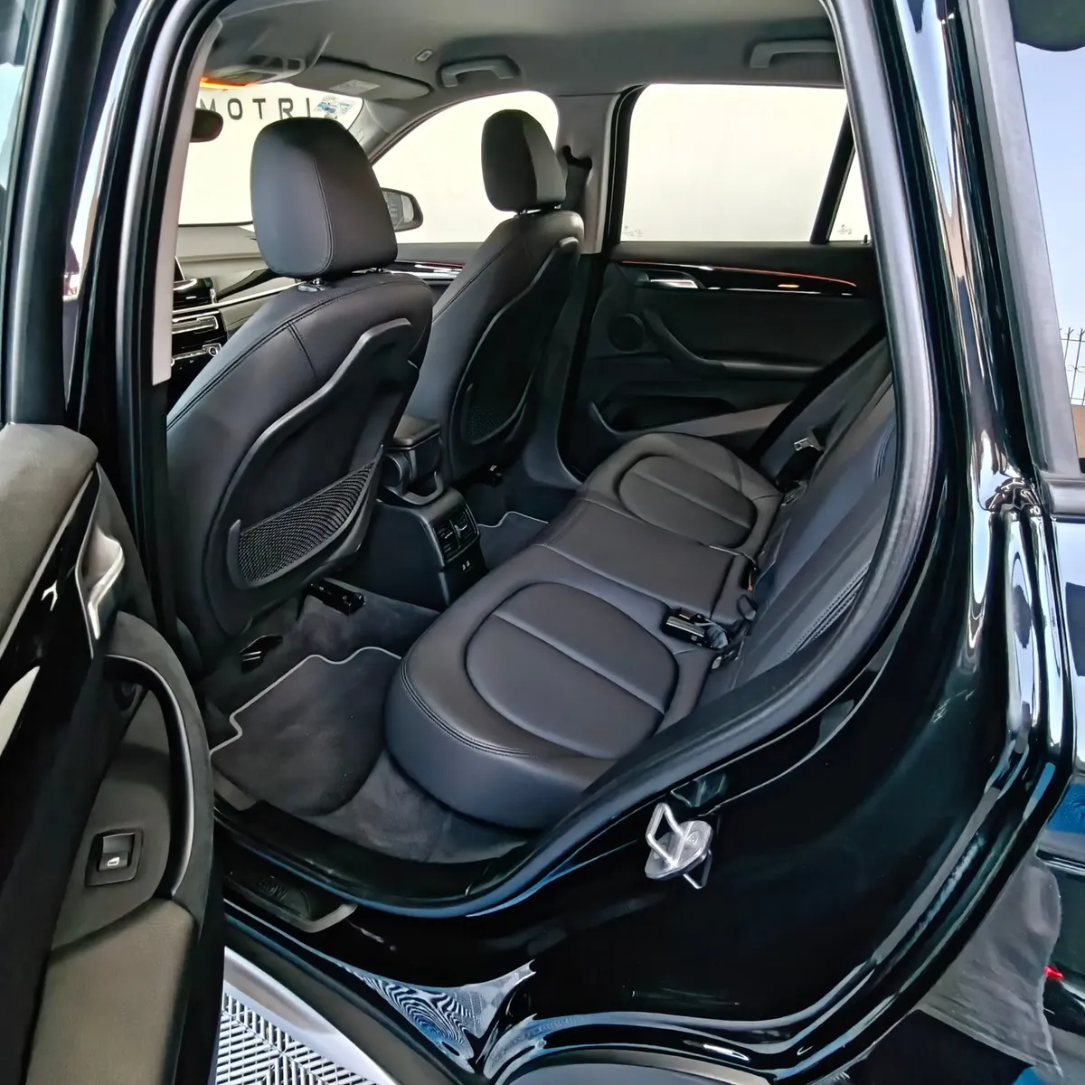 Vista interior desde la puerta trasera izquierda abierta que muestra la banca de asientos posteriores tapizados en piel negra, el espacio para piernas, las salidas de aire acondicionado y el respaldo del asiento del conductor de una camioneta SUV de lujo BMW X1 2.0 sDrive20i sDrive 20iA X Line transmisión automática año 2022 seminueva en ángulo oblicuo izquierdo.
