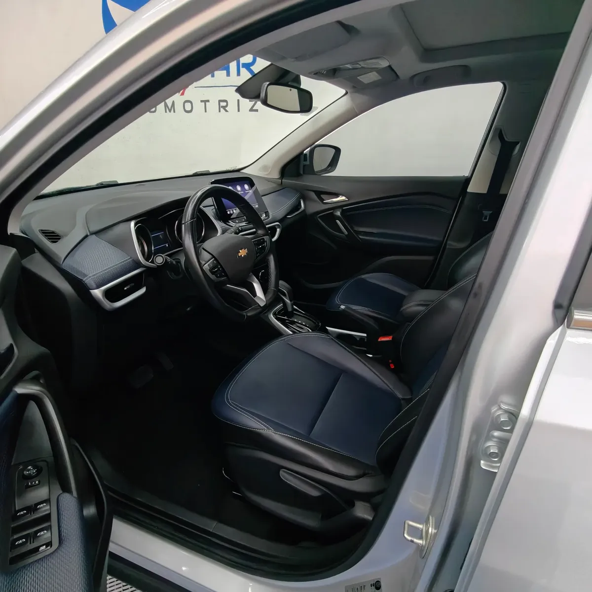 Vista interior desde la puerta del conductor abierta que muestra el puesto de mando con volante multifunción, el asiento delantero izquierdo forrado en piel bitono azul y negro, los controles de la puerta y el panel de instrumentos de una camioneta SUV Chevrolet Tracker 1.2 Premier Turbo transmisión automática año 2023 seminueva en ángulo oblicuo izquierdo.
