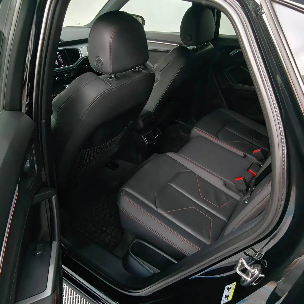 Vista interior desde la puerta trasera izquierda abierta que expone la banca de asientos posteriores tapizados en piel negra con costuras en rojo, los cinturones de seguridad de tres puntos y el espacio para piernas de una camioneta SUV Audi Q3 2.0 S Line 180 Hp transmisión DSG automática año 2022 seminueva en ángulo oblicuo izquierdo.