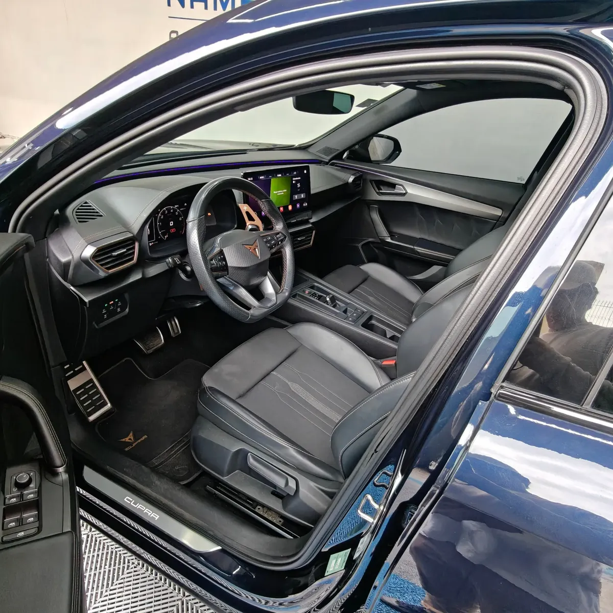 Vista interior desde la puerta del conductor abierta que muestra el puesto de mando, el asiento del piloto en piel y tela, el volante deportivo con paletas de cambio y los pedales de aluminio de una SUV Crossover deportiva Cupra Formentor 2.0 Mid 190 HP 2WD DSG7 año 2022 seminueva en ángulo oblicuo izquierdo.