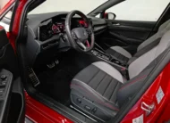 Vista interior desde la puerta del conductor abierta que muestra el puesto de mando, el asiento del piloto deportivo en tela bitono con ajuste eléctrico, el volante multifunción con acentos rojos, los pedales deportivos y la consola de un hatchback deportivo Volkswagen Golf GTI 2.0 241 HP DSG7 año 2024 seminuevo en ángulo oblicuo izquierdo.