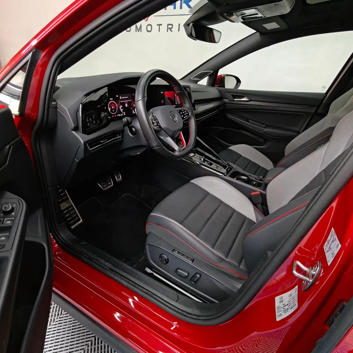 Vista interior desde la puerta del conductor abierta que muestra el puesto de mando, el asiento del piloto deportivo en tela bitono con ajuste eléctrico, el volante multifunción con acentos rojos, los pedales deportivos y la consola de un hatchback deportivo Volkswagen Golf GTI 2.0 241 HP DSG7 año 2024 seminuevo en ángulo oblicuo izquierdo.