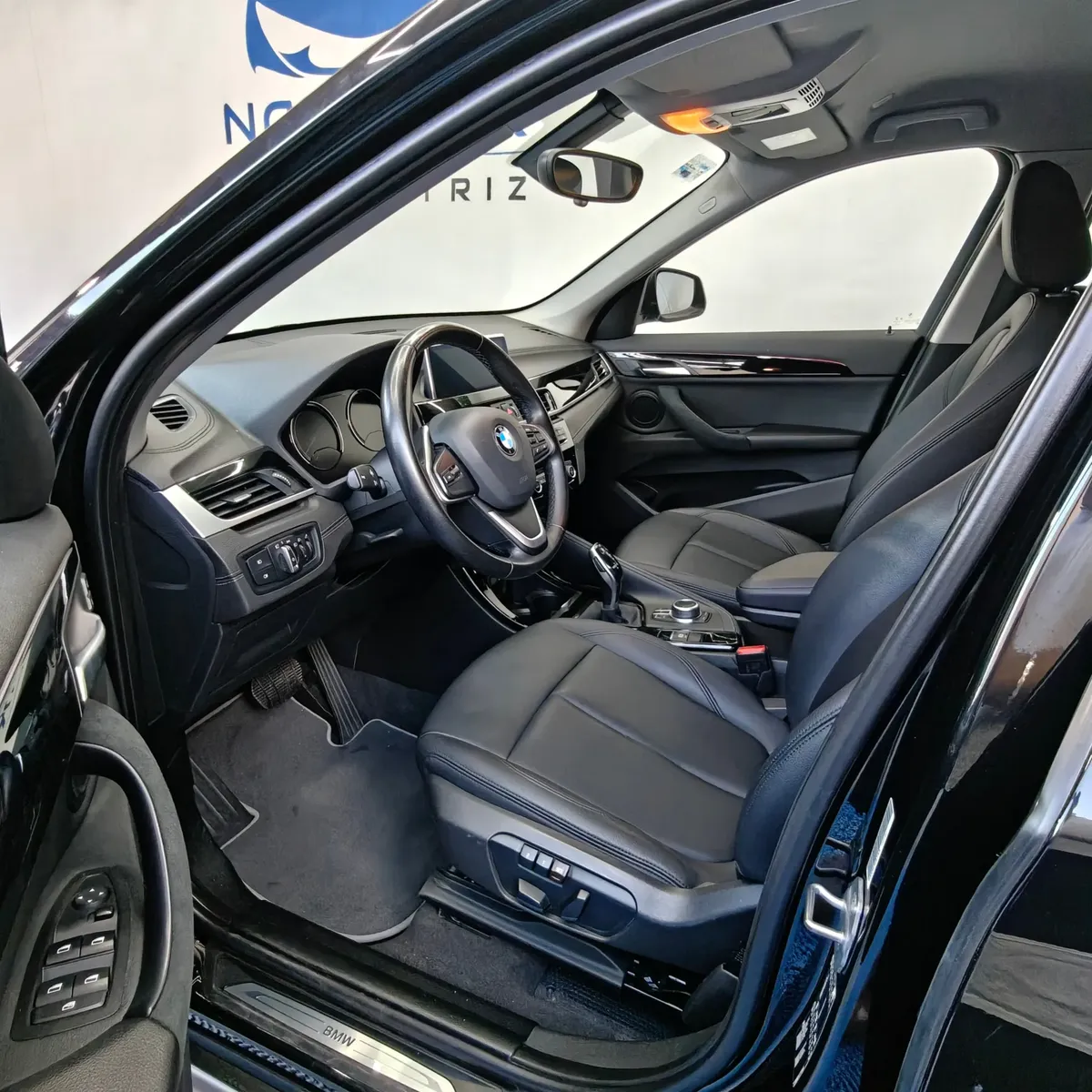 Vista interior desde la puerta del conductor abierta que muestra el puesto de mando, el asiento del piloto en piel negra con memoria y ajustes eléctricos, el volante multifunción con emblema BMW, la consola central y el tablero de una camioneta SUV de lujo BMW X1 2.0 sDrive20i sDrive 20iA X Line transmisión automática año 2022 seminueva en ángulo oblicuo izquierdo.