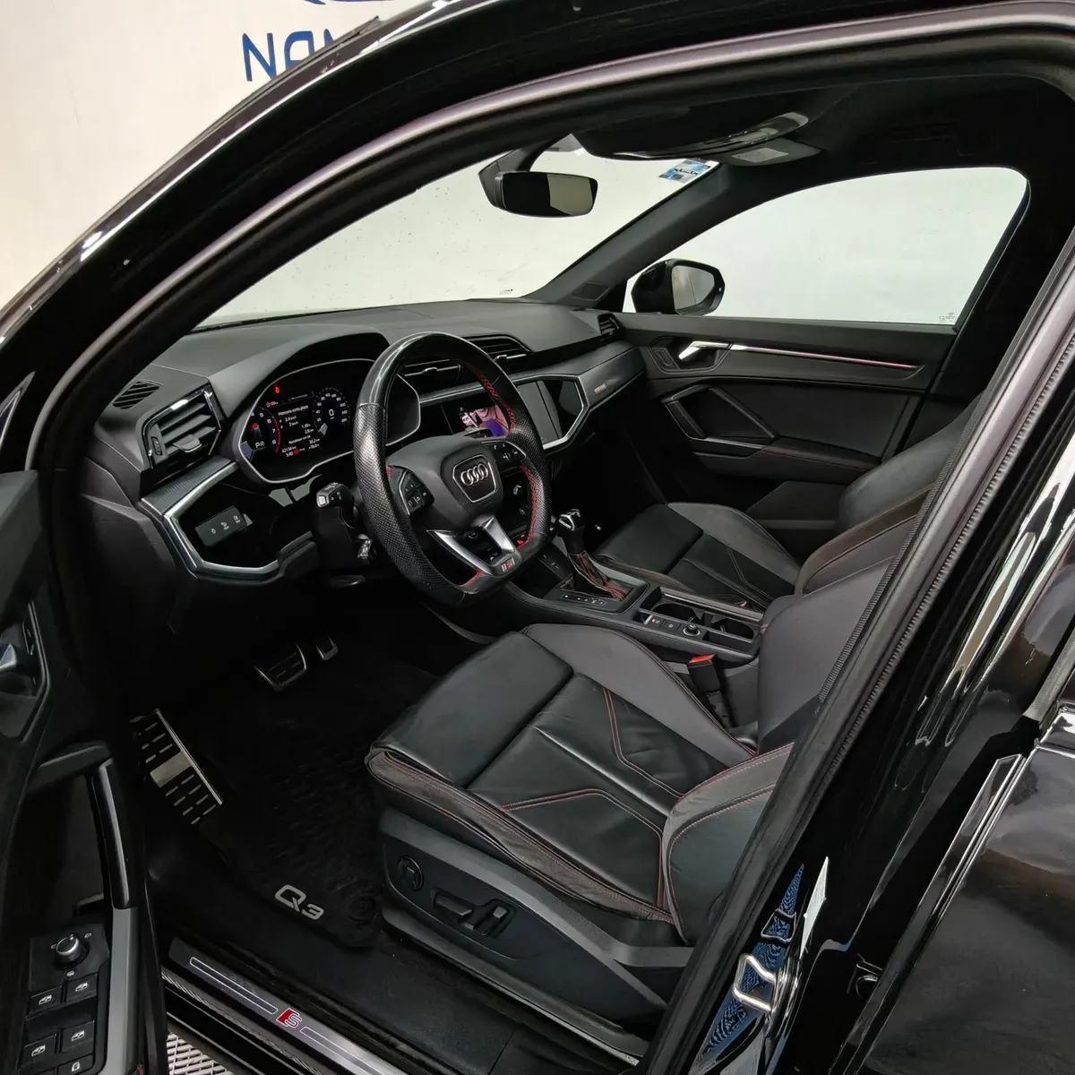 Vista interior desde la puerta del conductor abierta que muestra el puesto de mando ergonómico con volante deportivo achatado S Line, el asiento del piloto en piel negra con costuras rojas y ajustes eléctricos, pedales de aluminio y el cuadro digital Virtual Cockpit de una camioneta SUV Audi Q3 2.0 S Line 180 Hp transmisión DSG automática año 2022 seminueva en ángulo oblicuo izquierdo.