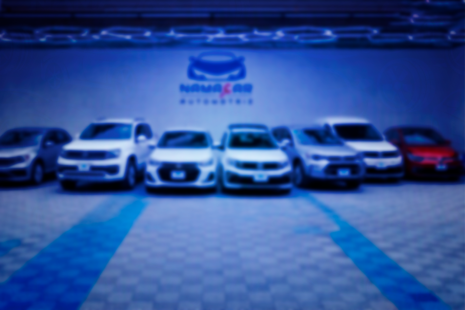 Vista frontal general y panorámica de la sala de exhibición de Namacar Automotriz, mostrando una variedad de autos seminuevos multimarca (sedanes, SUVs, pickups) alineados frente al logotipo de la agencia en una pared iluminada de azul, en Ciudad de México.